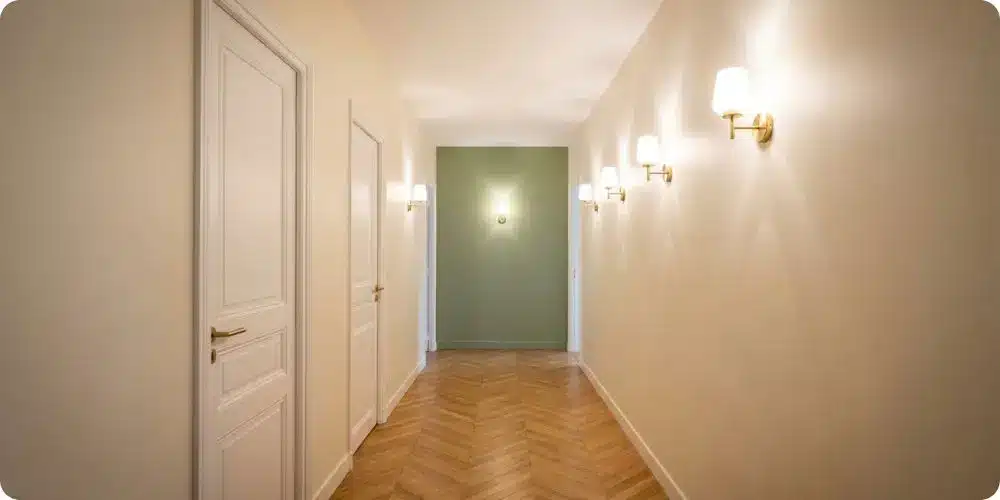 Couloir long avec murs latéraux crème et mur du fond peint en vert sauge, parquet chevrons, quatre appliques laiton créant un effet de profondeur
