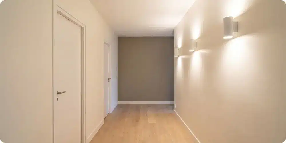Couloir sans fenêtre peint en blanc cassé avec trois appliques murales LED à 3 000 K et mur du fond greige créant un effet de profondeur