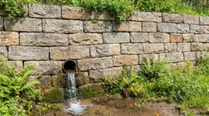 Une barbacane en tuyau évacuant de l'eau à travers un mur de soutènement en pierres sèches entouré de végétation.