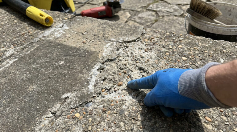 Une main gantée de bleu montre une zone de béton désactivé dégradé, avec des outils de réparation en arrière-plan.